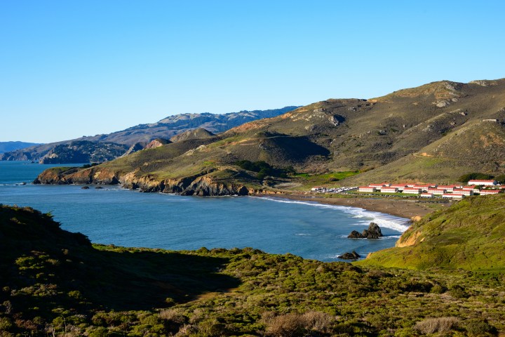 Coastal landscape with mountains, ocean bay, beach, and distant buildings under a clear blue sky.